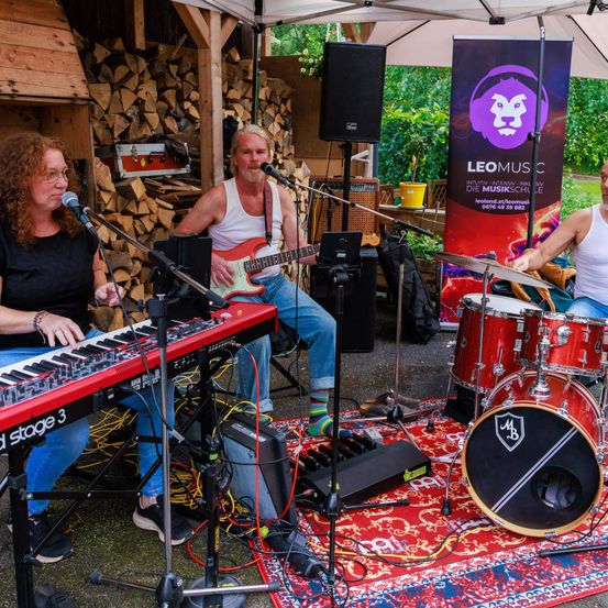 Drei Musiker spielen im Freien auf einer Bühne. Eine Frau spielt die Tastatur, ein Mann mit einer Gitarre steht und ein anderer Mann spielt die Trommeln. Dahinter befindet sich ein Banner und ein Lautsprecher.