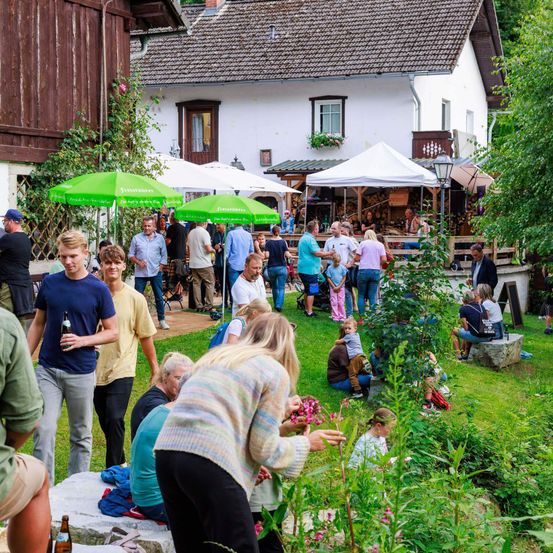 Eine Gruppe von Menschen versammelt sich in einem Gartenbereich im Freien, einige unter grünen Regenschirmen. Die Leute unterhalten sich und stehen in Gruppen. Eine Frau pflückt Blumen und ein Mann hält eine Flasche. Der Bereich hat ein rustikales Haus mit einer Veranda, und es gibt einen kleinen Garten mit Blumen davor.