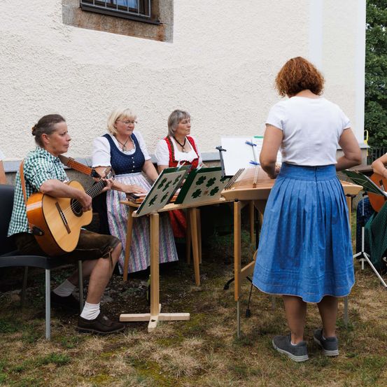 Fünf Personen sind im Freien versammelt, ein Mann spielt Gitarre, drei Frauen sitzen mit Notenblättern vor ihnen. Eine Frau in einem blauen Rock steht in der Nähe.