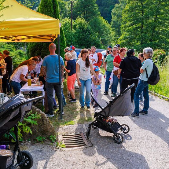 Eine Gruppe von Menschen versammelt sich im Freien, einige tragen Mützen und Turnschuhe. Eine Person schiebt einen Kinderwagen. Eine andere sitzt unter einem gelben Zelt. In der Nähe steht ein Tisch mit Essen darauf.
