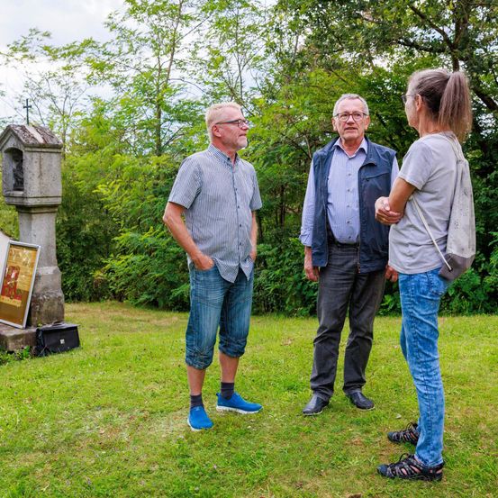 Drei Personen unterhalten sich im Freien, einer der Männer trägt eine Brille und blaue Schuhe, ein anderer Mann hält die Schulter einer Frau mit Sandalen. Hinter ihnen steht ein kleines Steingebäude mit einem Kreuz.