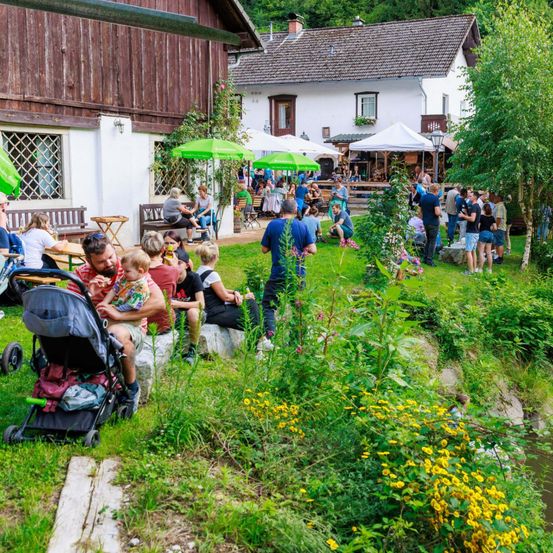 Menschen versammeln sich in einem Gartenbereich mit Häusern und einem Bach, einige unter Schirmen, andere in geselliger Runde, mit Bäumen und Blumen im Hintergrund.