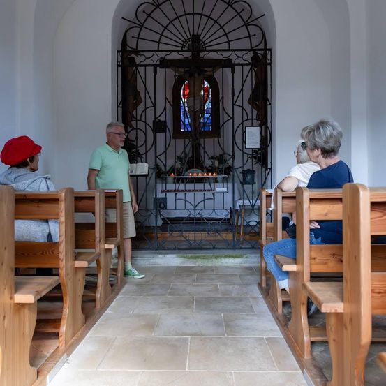 Ein Mann steht in einer Kirche und spricht zu einer Gruppe von Menschen, die auf hölzernen Kirchenbänken sitzen. Hinter ihm befindet sich ein schmiedeeisernes Tor mit einem Kruzifix und einem Buntglasfenster.