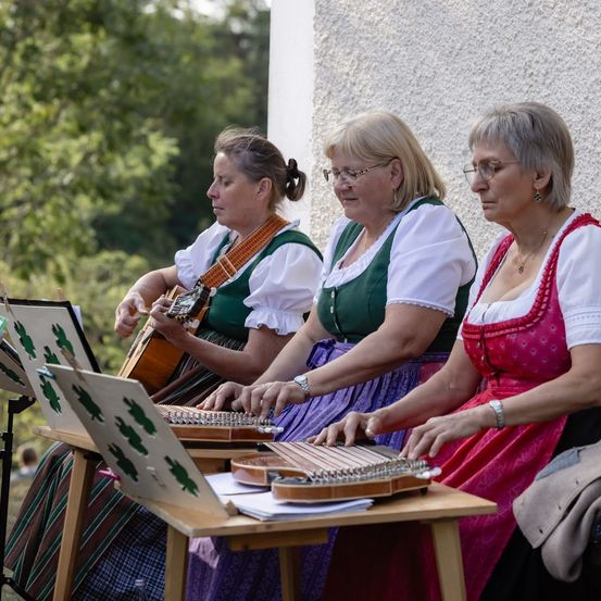 Drei Frauen in traditioneller Kleidung spielen Saiteninstrumente auf einem Holztisch im Freien.