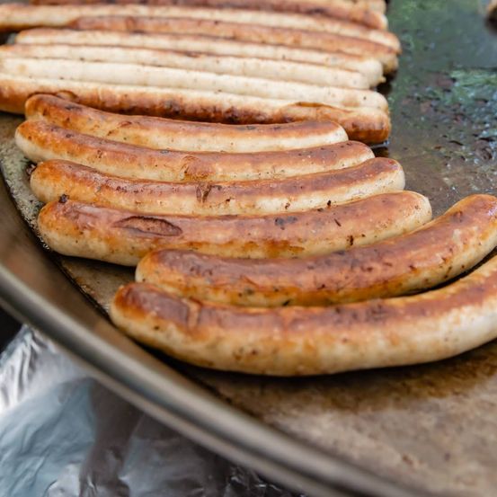 Eine Nahaufnahme von mehreren Würstchen, die auf einem Metalltablett gegrillt werden, mit einer goldbraunen Kruste und einem leicht verkohlten Aussehen.