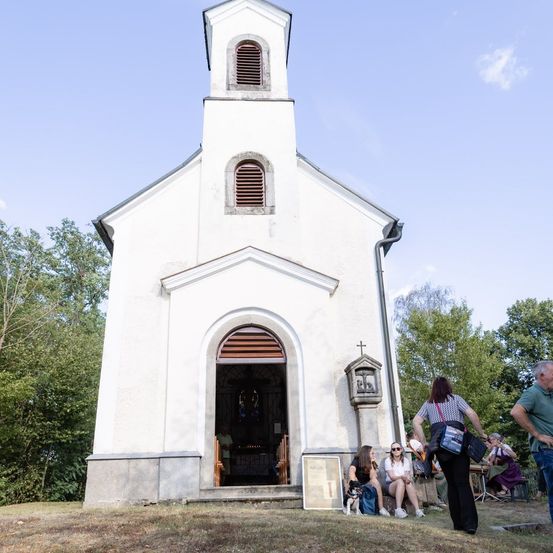 Eine kleine weiße Kirche mit einem Turm und einem gewölbten Eingang, umgeben von Grün. Mehrere Menschen sind draußen versammelt, einige sitzen und andere stehen.