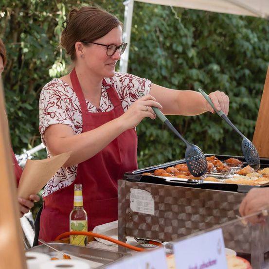 Eine Frau in einem roten Schürze kocht Essen auf einem Grill im Freien. Sie benutzt einen Kochlöffel, um das Essen zu wenden.