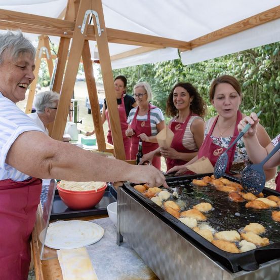 Eine Gruppe von Frauen in roten Schürzen kocht Essen auf einem Grill unter einem Zelt. Eine Frau lächelt und hält einen Schöpflöffel.