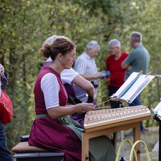 Eine Frau in einem gepunkteten Kleid spielt Zither, während einige Leute hinter ihr in einem Park stehen.