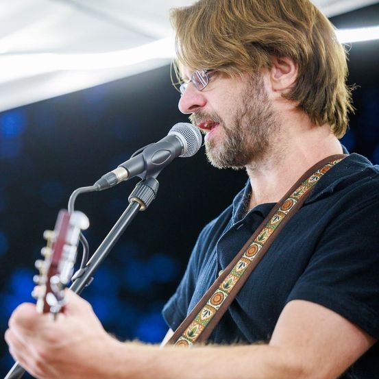 Ein Mann mit Brille und Bart singt in ein Mikrofon, während er eine Gitarre hält.