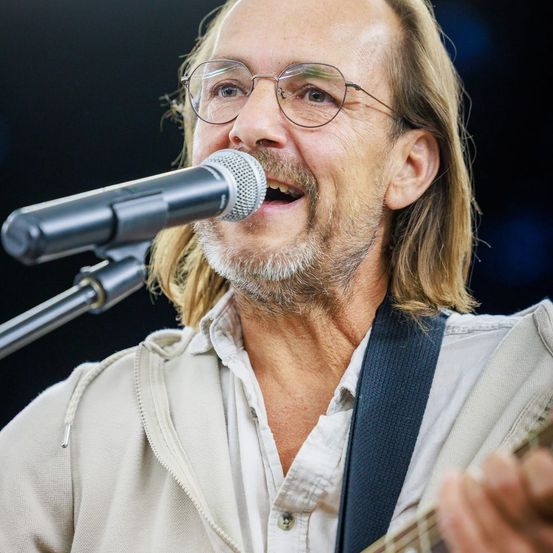 Ein Mann mit langen Haaren und einer Brille singt in ein Mikrofon, während er die Gitarre spielt.
