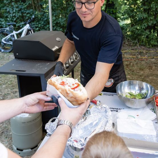 Ein Mann in einem blauen T-Shirt und schwarzen Handschuhen bereitet ein Sandwich im Freien zu, mit einem Grill und einer Schüssel Salat in der Nähe.