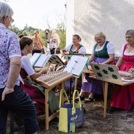 Eine Gruppe von Frauen in traditioneller Kleidung sitzt um Tische herum und spielt verschiedene Musikinstrumente. Ein Mann steht in der Nähe und beobachtet sie.