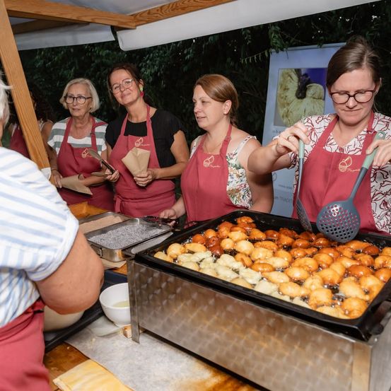 Fünf Frauen in roten Schürzen kochen im Freien. Eine Frau brät Essen in einer Pfanne, während andere zusehen. Ein Mann in einem gestreiften Hemd beobachtet von der Seite.