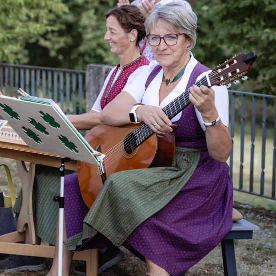 Zwei Frauen sitzen auf einer Parkbank, eine spielt Gitarre, während die andere Musiknoten liest.