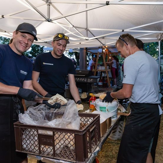 Drei Männer in Schürzen und Handschuhen bereiten Essen unter einem Zelt zu, mit einem Grill und Eimern in der Nähe.