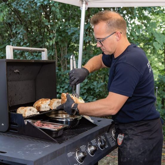 Ein Mann in einem blauen Shirt und einer schwarzen Schürze grillt im Freien auf einem Grill.