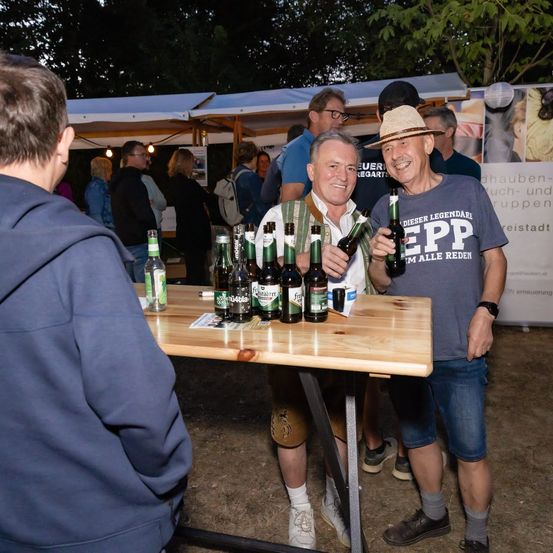 Zwei Männer stehen neben einem Tisch voller Bierflaschen und Dosen und lächeln für ein Foto bei einer Veranstaltung im Freien.