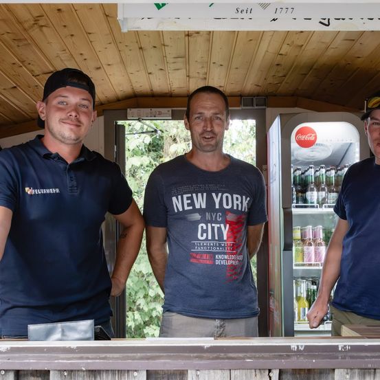 Drei Männer stehen vor einer Holzstruktur und lächeln für ein Foto. Der Mann in der Mitte trägt ein 'New York City' T-Shirt. Dahinter ist ein Kühlschrank mit Getränken zu sehen.