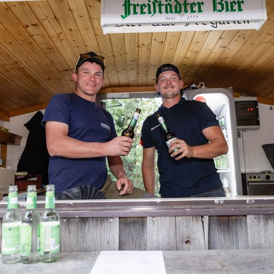 Zwei Männer in blauen Shirts stehen hinter einer Bartheke, jeder hält eine Flasche Freitadter Bier, mit einer hölzernen Decke und einem Schild über ihnen.
