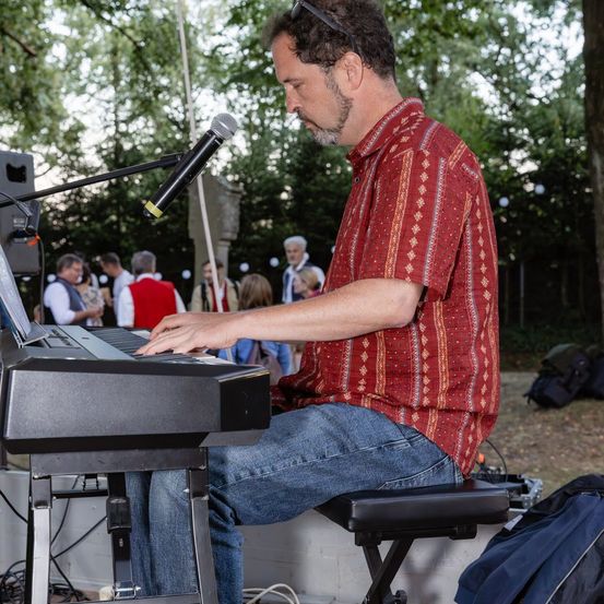 Ein Mann spielt eine Tastatur in einer Außenumgebung, mit einem Mikrofon und Lautsprechern in der Nähe. Er trägt ein rotes Hemd und Jeans. Im Hintergrund stehen mehrere Menschen und unterhalten sich.