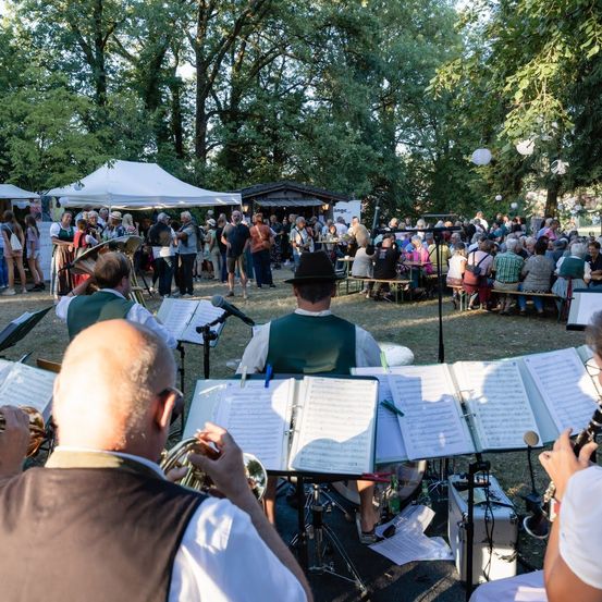 Eine Gruppe von Musikern spielt im Freien, während eine Menschenmenge zusieht. Die Musiker sitzen mit Musikinstrumenten und Notenblättern. Das Publikum sitzt auf Bänken und Tischen unter einem Zelt.