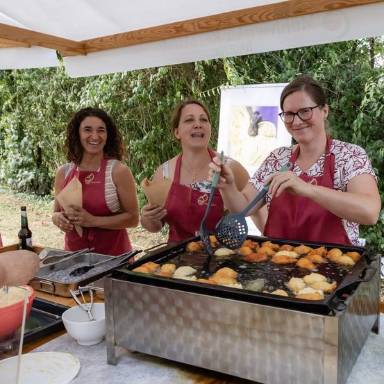 Drei Frauen in roten Schürzen kochen im Freien, eine frittiert Essen in einer Pfanne, eine andere hält einen Schöpflöffel und die dritte hält einen Papierkegel.