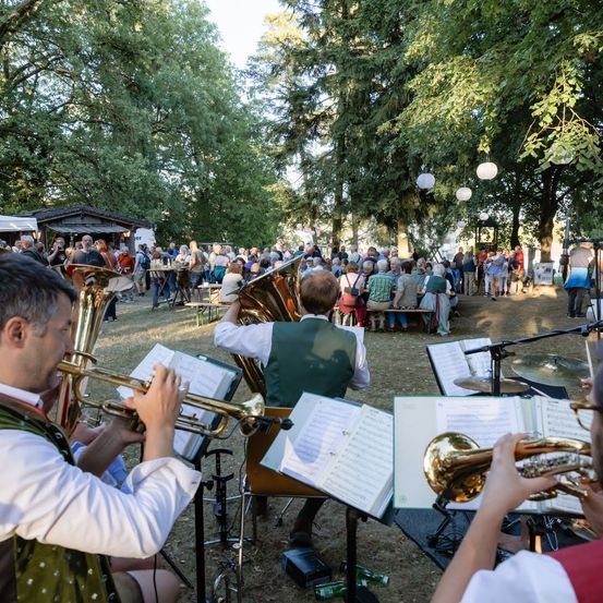 Eine Blechbläsergruppe spielt im Freien in einem Park, während eine Menge Menschen zuschaut. Die Musiker sitzen und spielen verschiedene Instrumente, darunter Trompeten und Tuben.