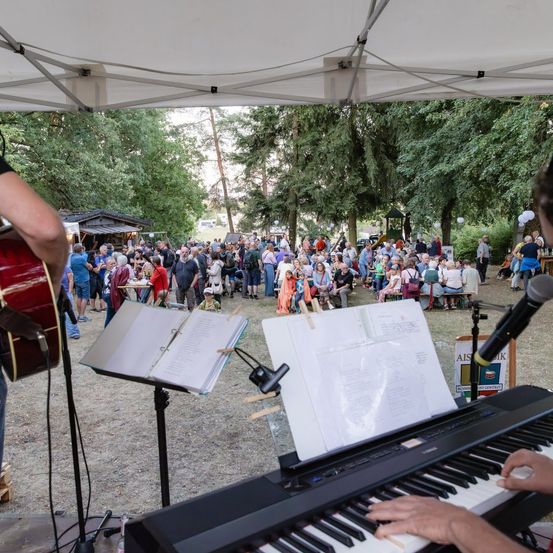 Eine Band spielt auf der Bühne unter einem Zelt, mit einem Pianisten und Gitarristen, während sich eine Menge versammelt.