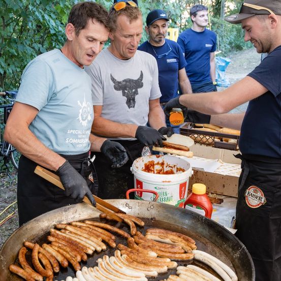 Eine Gruppe von Männern kocht Würste in einer großen Pfanne im Freien. Ein Mann fügt den Würsten Sauce hinzu, während andere sie vorbereiten.