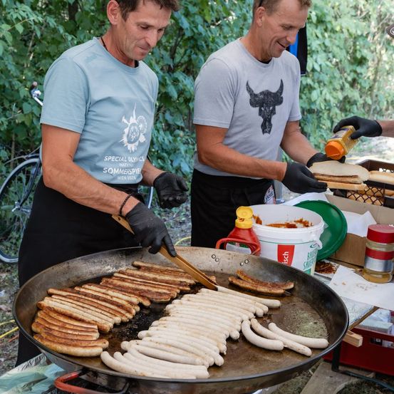Zwei Männer kochen Würstchen in einer großen Bratpfanne im Freien. Ein Mann wendet die Würstchen mit einer Zange, während der andere ein Sandwich vorbereitet. Im Hintergrund sind verschiedene Gewürze und ein Fahrrad zu sehen.