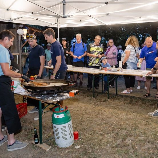 Eine Gruppe von Menschen versammelt sich um einen Tisch unter einem Zelt, wo zwei Männer auf einer großen Pfanne kochen. Der Tisch ist mit Flaschen, Tassen und anderen Gegenständen bestückt. Menschen stehen und schauen zu, einige tragen Hüte und Turnschuhe.