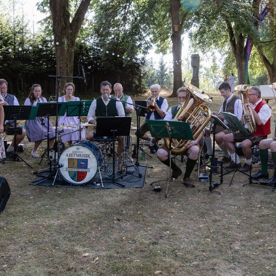 Eine Gruppe Musiker in traditioneller Kleidung spielt im Freien, mit Instrumenten wie Tubas und Trommeln auf einer Grasfläche aufgestellt.