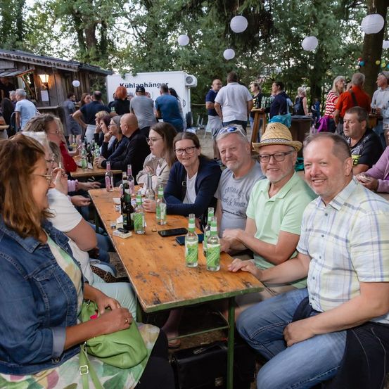 Eine Gruppe von Menschen sitzt an einem Tisch in einer Außenumgebung, mit mehreren Flaschen und einem Handy auf dem Tisch. Sie lächeln und scheinen ihre Zeit miteinander zu genießen. Im Hintergrund befinden sich Bäume und ein Gebäude.