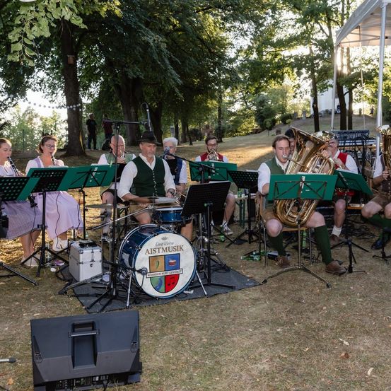 Eine Gruppe von Musikern in traditioneller Kleidung spielt im Freien, mit einem Schlagzeug und Blechblasinstrumenten auf dem Boden aufgestellt.