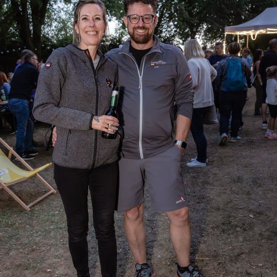 Ein Mann und eine Frau stehen eng beieinander und lächeln, mit einer Flasche in der Hand der Frau. Sie sind in Freizeitkleidung gekleidet. Hinter ihnen geht eine Gruppe von Menschen in einem staubigen Bereich mit Zelten und Bäumen.