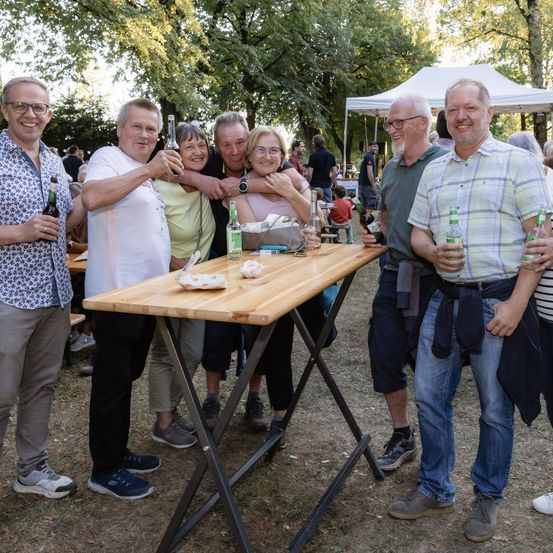 Eine Gruppe Erwachsener, die in einem Park lächelnd für ein Foto posiert, mit einem Tisch und Getränken vor ihnen.