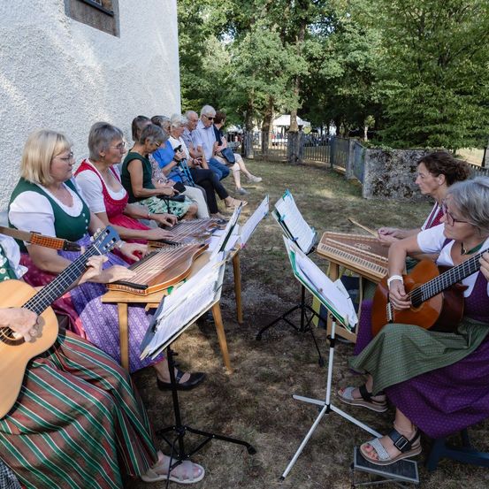 Eine Gruppe älterer Frauen spielt Gitarren und andere Instrumente im Freien, mit einem Gebäude und Bäumen im Hintergrund.