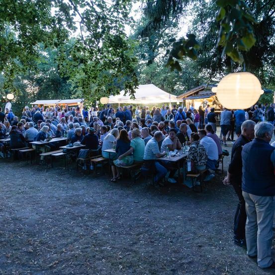 Eine große Gruppe von Menschen ist für eine Veranstaltung im Freien versammelt, sitzt an Tischen unter Zelten mit Laternen. Bäume und Grünflächen umgeben das Gebiet.
