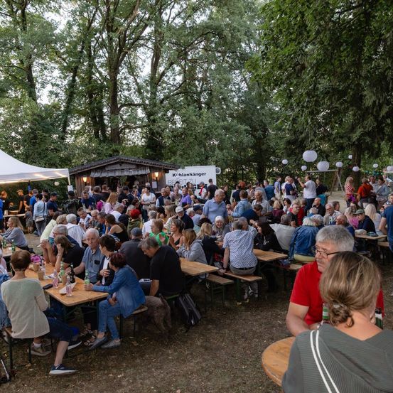 Eine große Gruppe von Menschen ist zu einer sozialen Veranstaltung in einem Park versammelt. Tische und Bänke sind aufgestellt, und einige Leute stehen und unterhalten sich. Bäume und ein Zelt bieten Schatten.