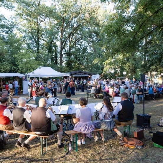 Eine Band spielt Musik im Freien in einem Park, wo eine Menschenmenge auf Bänken und Tischen sitzt, einige in traditioneller Kleidung, unter einem Zelt und Bäumen.