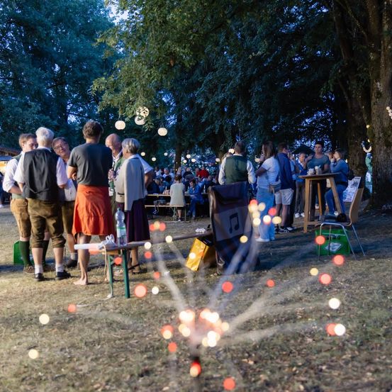Eine Gruppe von Menschen versammelt sich nachts in einem Park, einige stehen und andere sitzen um Tische herum, mit Bäumen und hängenden Lichtern im Hintergrund.