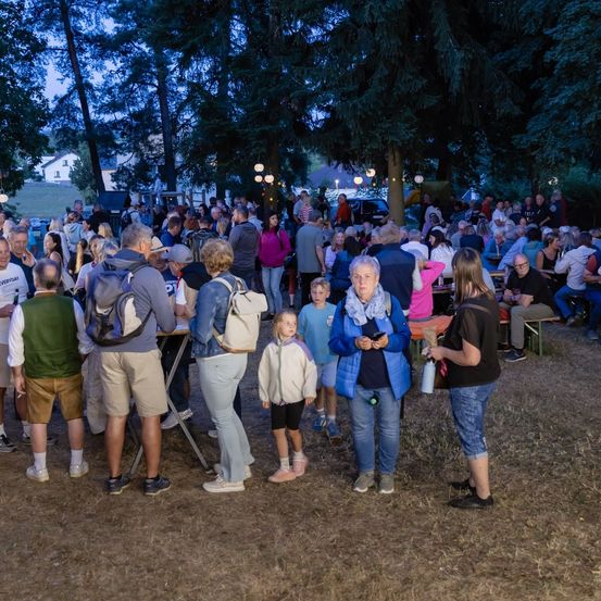 Eine große Gruppe von Menschen versammelt sich bei Nacht im Freien. Einige stehen, andere sitzen auf Bänken. Sie sind von Bäumen und einigen Straßenlaternen umgeben.
