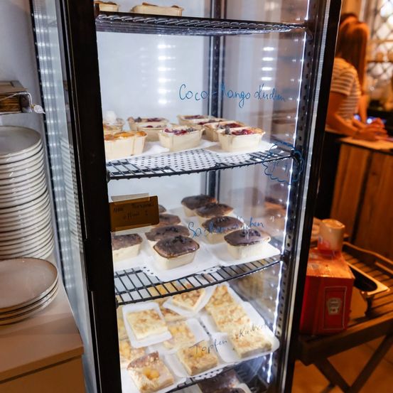 Eine Vitrine mit verschiedenen Gebäckstücken, darunter Törtchen und Stücke, mit handgeschriebenen Etiketten. Eine Person steht hinter der Vitrine, und ein Stapel Teller ist in der Nähe.