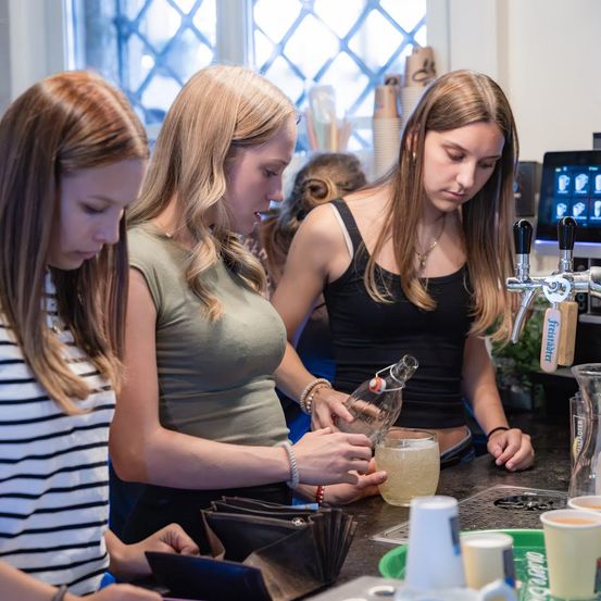 Drei junge Frauen stehen an einer Bartheke, eine gießt ein Getränk in ein Glas, während die anderen zusehen. Die Theke ist mit Tassen, Flaschen und einer Kasse übersät.