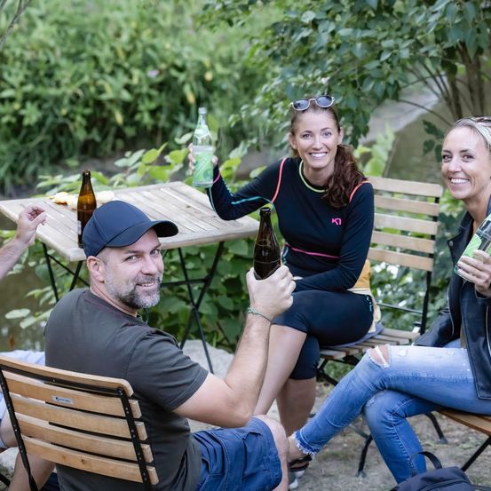 Vier Personen sitzen um einen Tisch in einem Garten, lächeln und halten Flaschen. Ein Mann mit einer Kappe hält eine Flasche in der Hand. Eine Frau mit Sonnenbrille auf dem Kopf sitzt neben ihm.