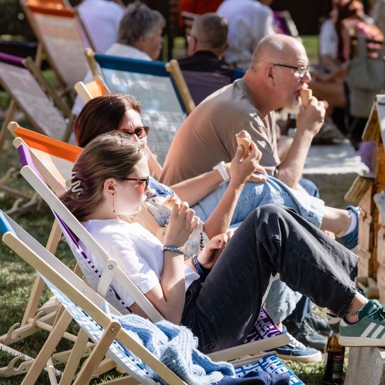 Eine Familie mit drei Personen, darunter ein junges Mädchen, eine Frau und ein Mann, sitzen auf Klappstühlen und essen Eis an einem sonnigen Tag.
