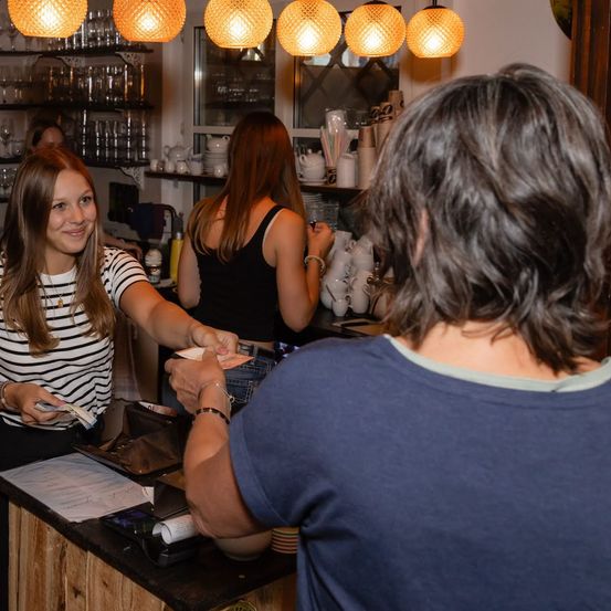 Eine Frau in einem gestreiften Shirt lächelt und reicht einer anderen Frau Geld an einem Tresen in einem Café.