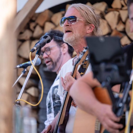 Drei Männer treten auf der Bühne auf. Der Mann in der Mitte singt in ein Mikrofon und hält eine Gitarre. Der Mann auf seiner linken Seite singt ebenfalls und spielt Gitarre. Der Mann auf seiner rechten Seite spielt Gitarre.