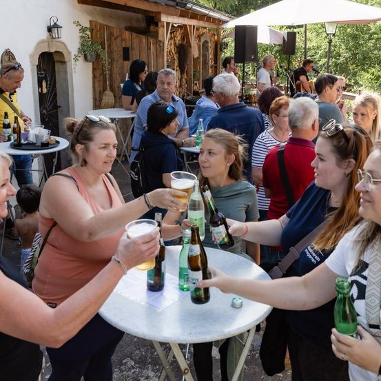Eine Gruppe von Menschen versammelt sich um einen Tisch im Freien und stößt mit Biergläsern an. Die Umgebung scheint ein zwangloses Outdoor-Event mit Tischen, Stühlen und einer Holzstruktur im Hintergrund zu sein.